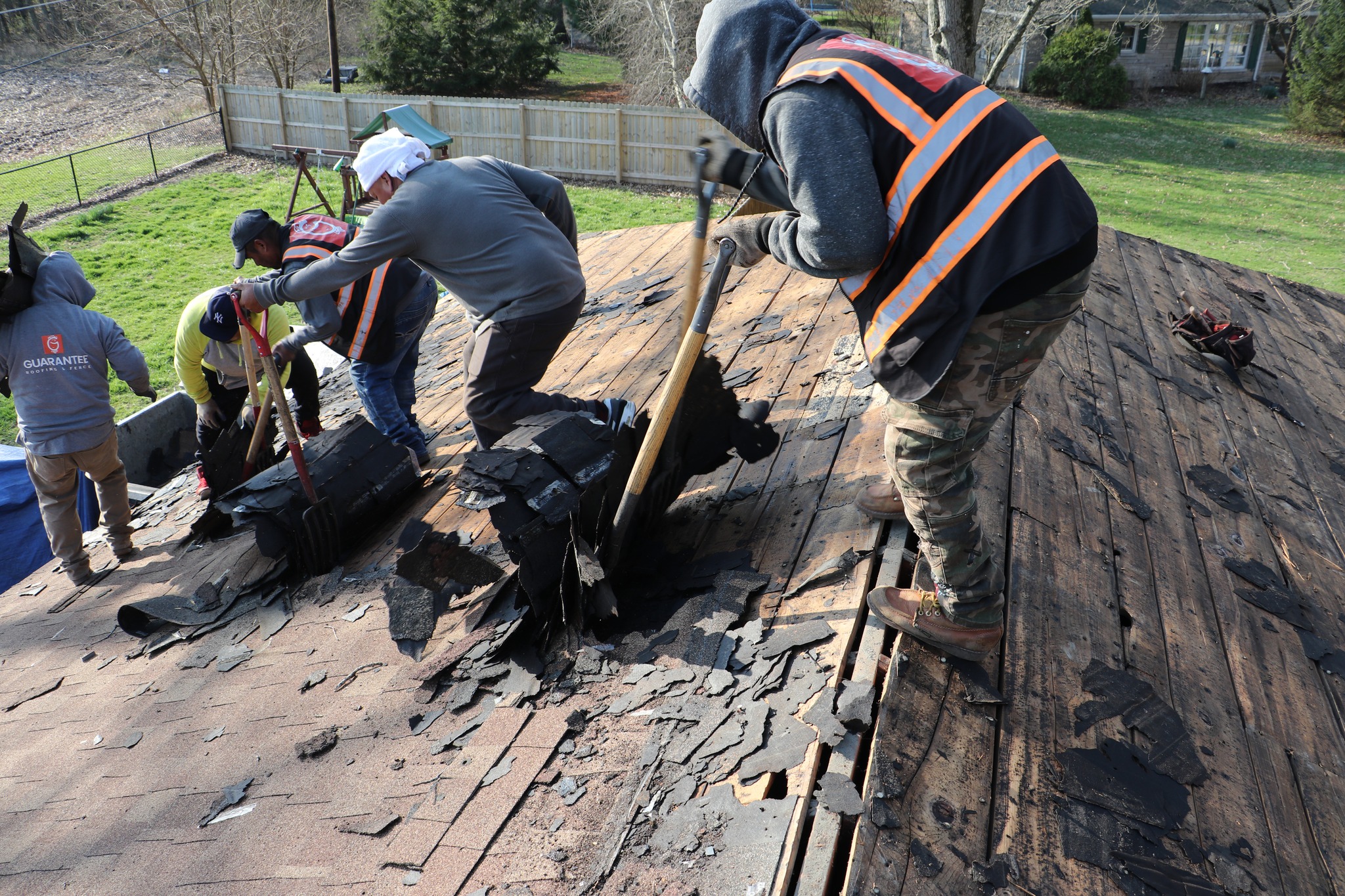 Guarantee Roofing roofers removing an old, worn roof before putting on a new one. The employees are wearing safety gear and using shovels to scrape away the old shingles. 