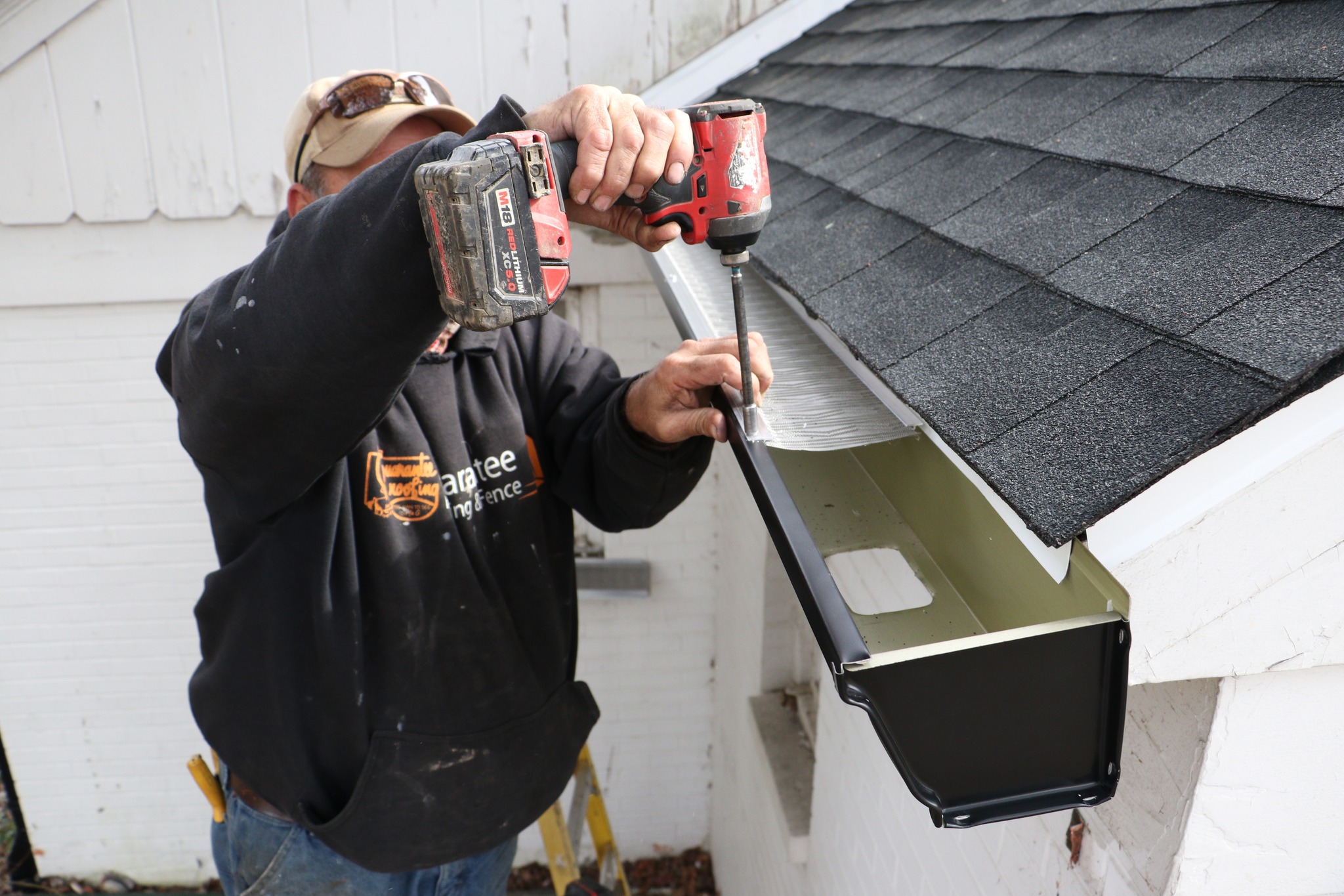 Installing New Gutters and Gutter Guards at a black and white home. There is a man in a dark hoodie using a power tool to screw in the gutter.