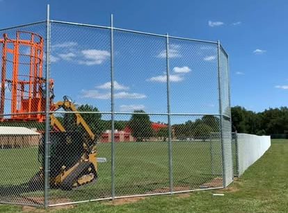Image of a large, silver, chainlink fence being installed for a baseball diamond backstop by Guarantee Roofing and Fence in Terre Haute Indiana. The yellow and orange lift is behind it, showing how the installer gets up there to do the taller parts.