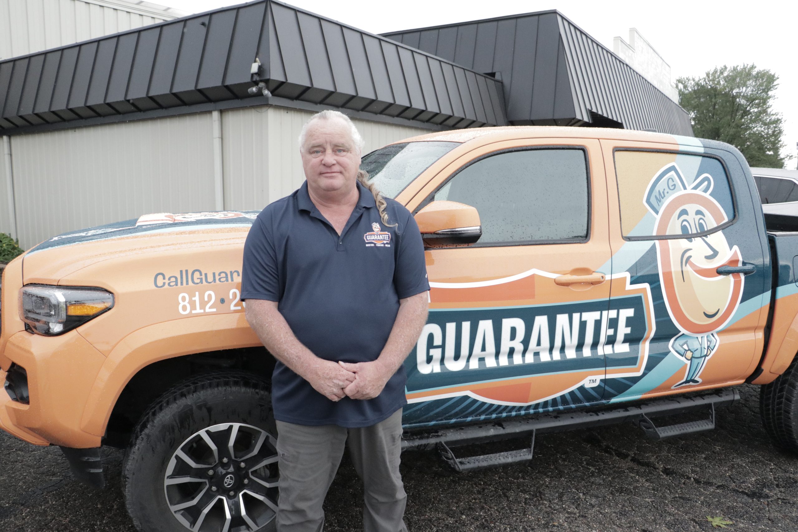 Mike Byrd, professional insurance company fighter, posing in front of his Guarantee Roofing and Fence work truck. He's an older white guy with a white ponytail and a soft smile.