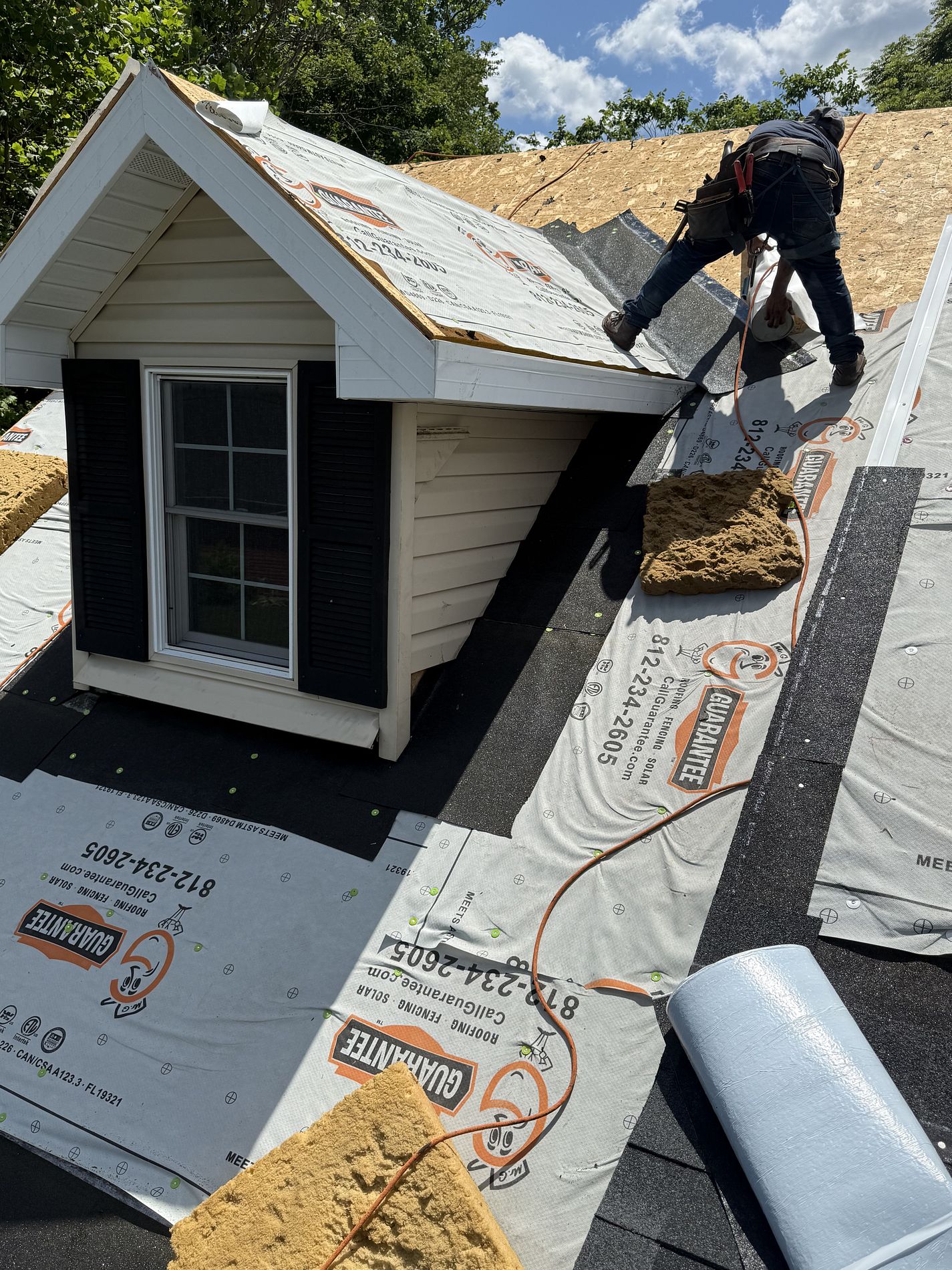Guarantee Roofing and Fence Company employee installing a new roof in Terre Haute. The man is perched on top of a gable, with Guarantee branded orange and black fabric below him. There is a small foam cushion off to the side. The couple of shingles that are installed around the edges are a dark grey color.