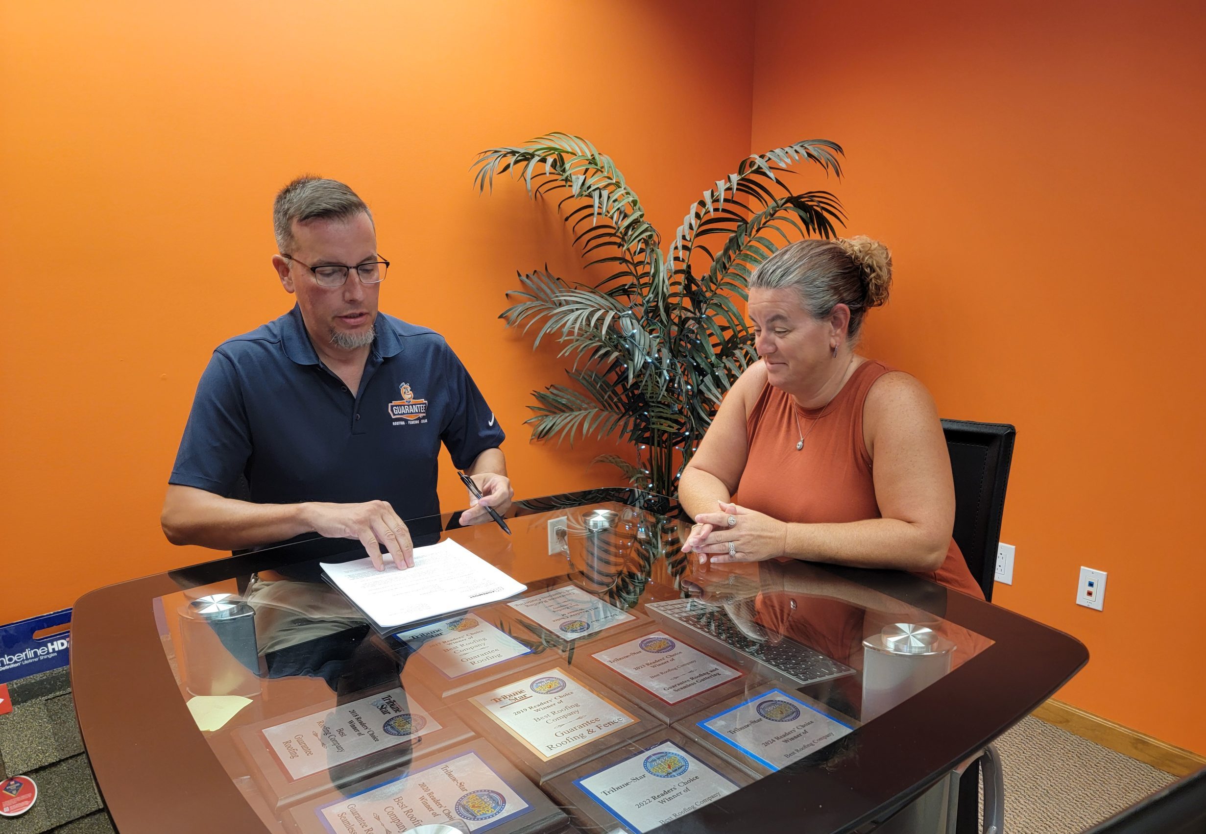 Middle-Aged white man in navy polo is a sales guy talking to a middle-aged white woman at a clear-top table. The walls are bright orange and they are looking at paperwork while talking.