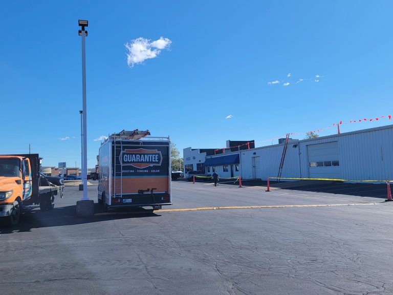 Guarantee Roofing and Fence is replacing the roof at Mace Ford, a local car dealership. There is a ladder on the side of the large, white building, with lines of orange flags draped near the edge of the roof. There is one little, bright white cloud in the otherwise beautiful blue sky.